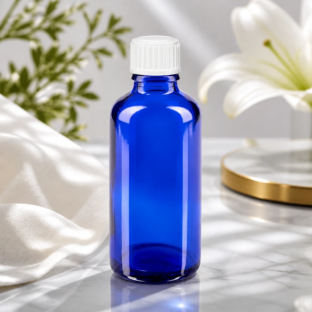 Flacon de pharmacie bleu 50ml bouchon vissable blanc standard Flacon en verre bleu de 50 ml muni d'un bouchon à vis blanc, posé à côté d'un lys blanc sur une surface en marbre