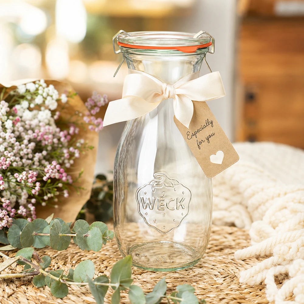 WECK Bouteille de jus de fruit de 530 ml RR60 décorée avec un ruban blanc et une étiquette cadeau arrangée entre des fleurs