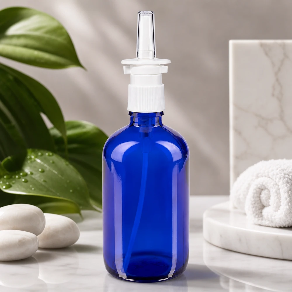Flacon de pharmacie bleu 100ml vaporisateur nasal blanc standard Flacon en verre bleu cobalt de 100 ml muni d'un embout vaporisateur, posé sur du marbre blanc à côté de pierres de bien-être