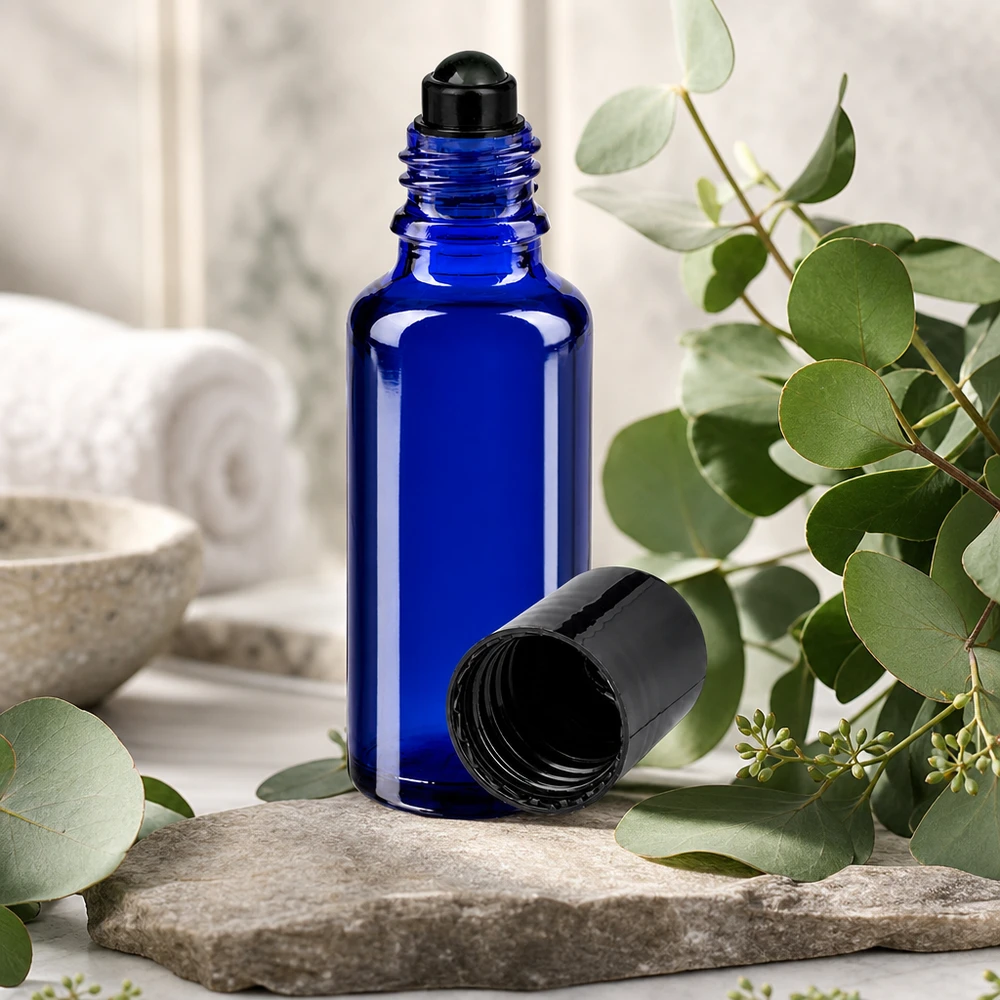 Flacon de déodorant en verre bleu 30 ml, déo à bille vide Flacon en verre bleu cobalt de 30 ml muni d'un bouchon à bille noir posé sur une pierre naturelle, entre des branches d'eucalyptus