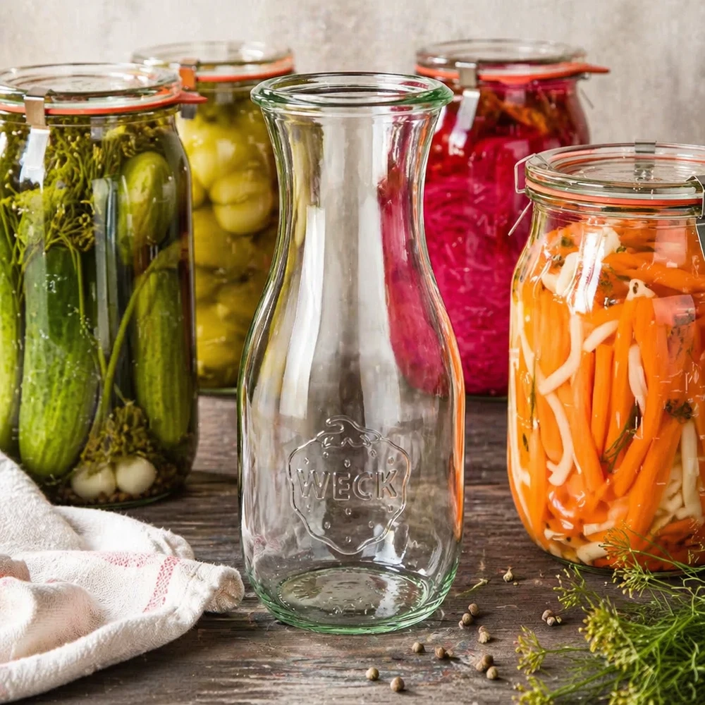 Bouteille à jus WECK RR60 de 530 ml Bouteille de jus WECK RR60 de 530 ml entourée de bocaux de légumes colorés et d'accessoires de conservation sur un fond en bois.