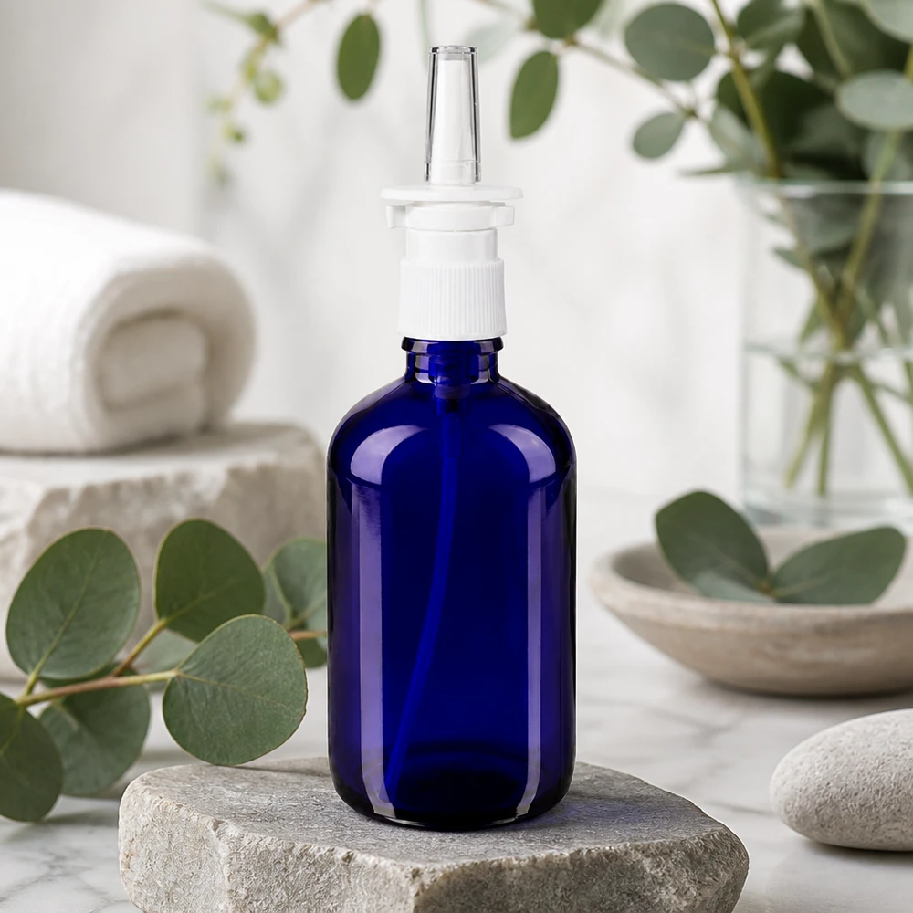 Flacon de pharmacie bleu 100ml vaporisateur nasal blanc standard Flacon en verre d'apothicaire bleu de 100 ml avec embout vaporisateur nasal, posé sur une pierre naturelle devant un eucalyptus