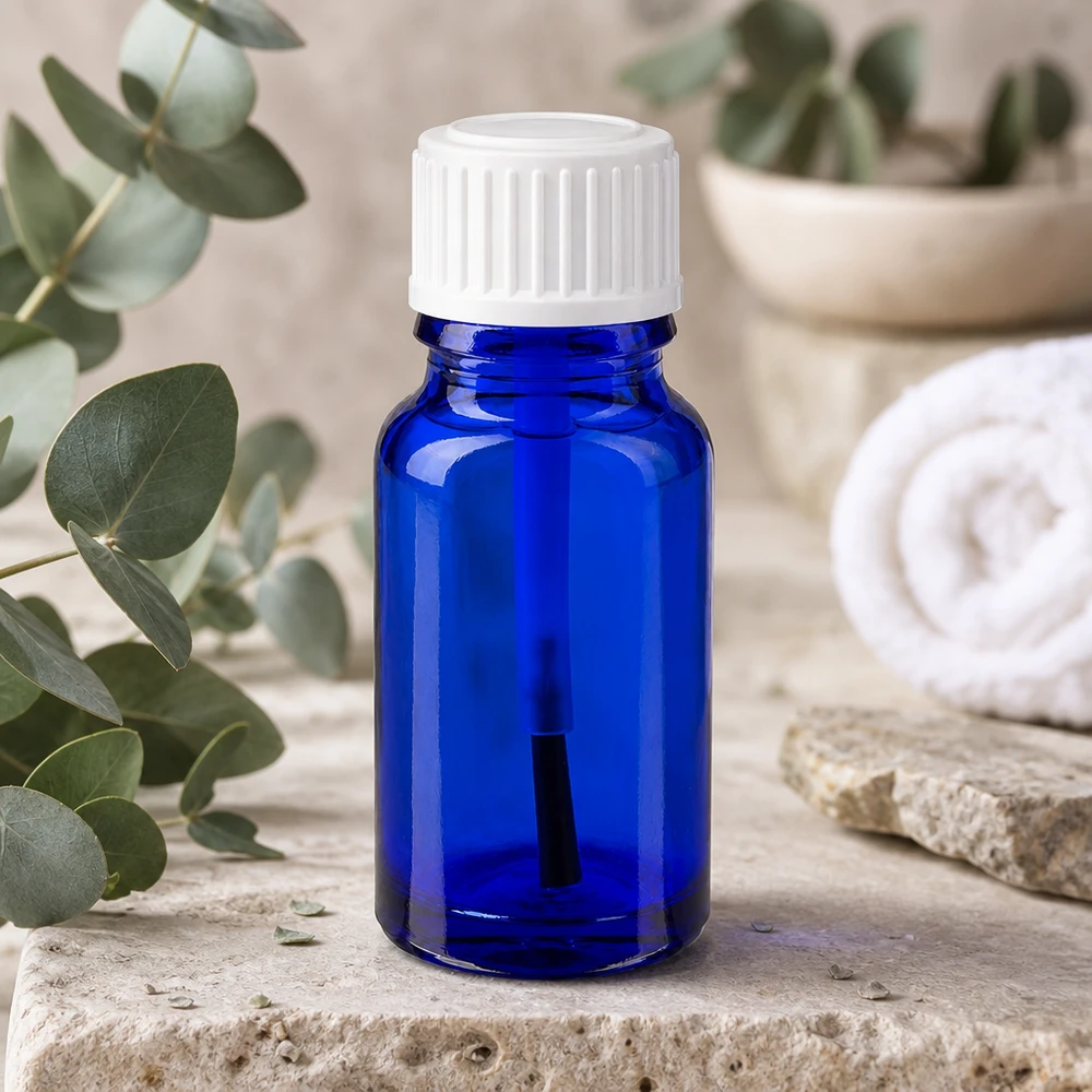 Flacon en verre bleu de 10 ml muni d'un embout pinceau blanc, posé à côté de branches d'eucalyptus sur une pierre naturelle